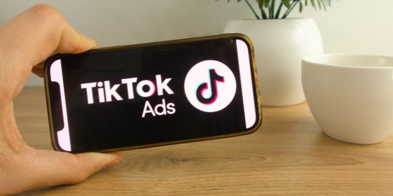 Uma mão segura um celular que mostra uma imagem com fundo preto e com o logo do TikTok Ads em destaque. Ao fundo há uma planta decorativa.