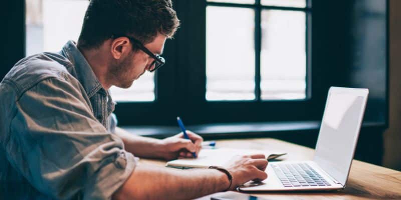 A imagem mostra um homem sentado em uma mesa, na frente dele está um notebook e uma folha de papel onde escreve.