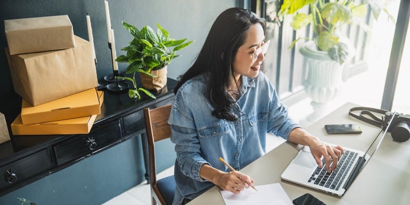 Uma mulher alegre trabalha em frente a um notebook enquanto faz anotações em uma folha de papel