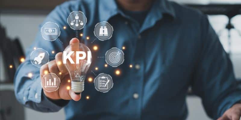 Um homem usando camisa azul segura uma lâmpada que está escrita KPI em efeito luminoso e ao redor do objeto, aparecem outras representações luminosas das vertentes do KPI
