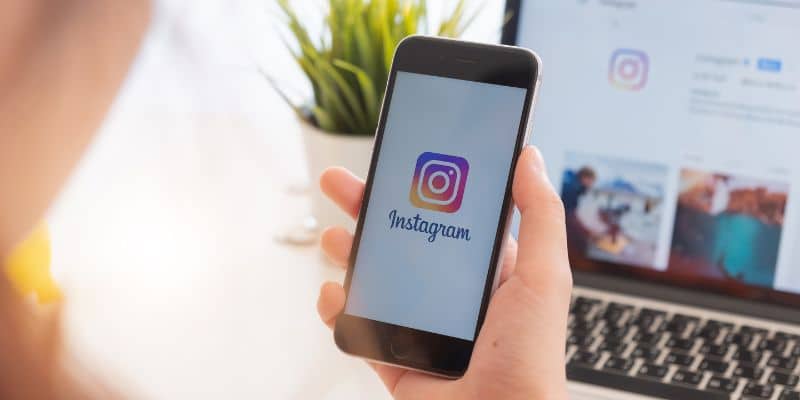 Imagem mostra uma pessoas usando o celular, na tela está o logo do Instagram. Ao fundo tem um notebook com a tela ligada em uma página do Instagram.