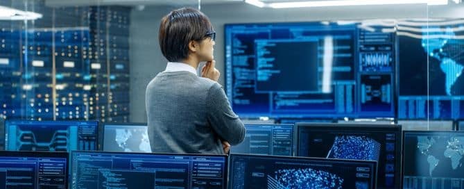 A imagem mostra um homem no centro, ele olha de forma concentrada para as telas de diversos computadores. Nas telas dos computadores está várias linhas de códigos.