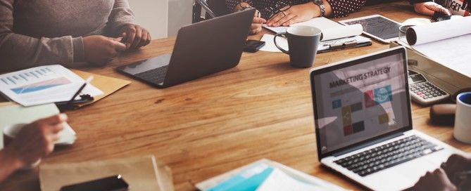 Em uma mesa de reunião há uma caneca, computadores e cadernos ao redor de algumas pessoas sentadas de frente uma para as outras, enquanto conversam