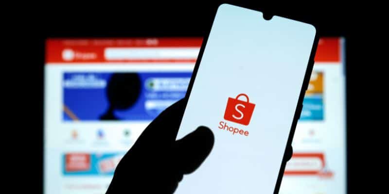 A imagem mostra uma pessoa segurando um celular, na tela do aparelho está a logo da Shopee. Logo atrás tem uma tela de computador com a página de compras inicial da Shopee.