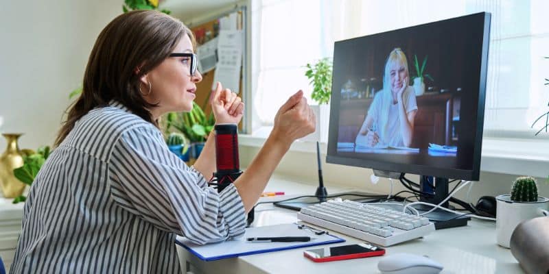 Uma mulher tem uma sessão de terapia via chamada de vídeo. Ela está sentada em uma cadeira em frente a um monitor no qual conversa com a paciente.