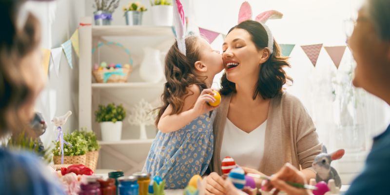 A imagem mostra uma mãe e uma filha comemorando a Páscoa com vários ovos coloridos em volta. A menina beija a bochecha da sua mãe que dá risada