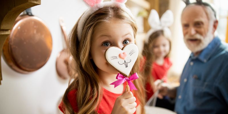 A imagem mostra uma garotinha sorrindo para a câmera, mas metade do seu rosto está escondido por um pirulito de chocolate em formato de coração e com o desenho de um focinho de coelho