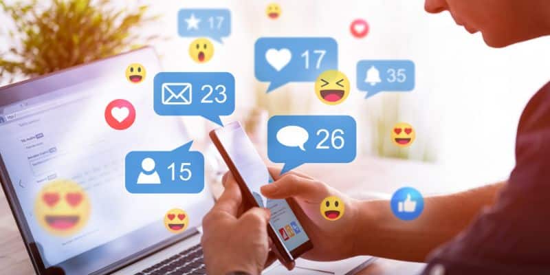 Uma pessoa mexe em um computador e há animações coloridas de ícones de redes sociais como emojis e notificações com likes, comentários e compartilhamentos