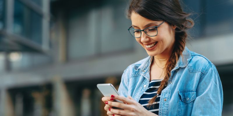 A imagem mostra uma moça de cabelo preso e jaqueta jeans mexendo no celular.