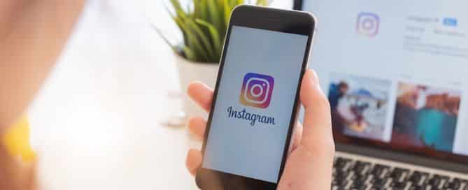 A imagem mostra em foco a mão de um homem que está segurando um celular. Na tela do aparelho está o logo do Instagram.