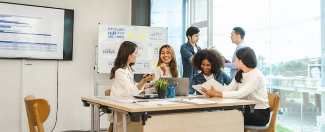 Um grupo de pessoas está reunida em uma empresa. No fundo há um quadro branco com anotações e há uma mesa em primeiro plano com computadores e objetos de escritório.
