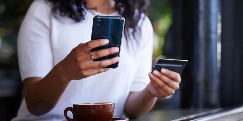 Mulher sentada em uma mesa ao ar livre com um celular na mão direita e um cartão de crédito na mão esquerda