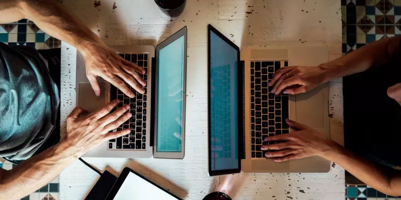 Duas pessoas, um homem e uma mulher, escrevem em um notebook, uma de frente para a outra em uma mesa. Vistos de cima é possível ver, além dos computadores, os objetos jogados em cima da mesa, como celulares