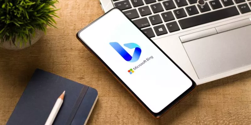 SEO para Bing no mobile A imagem mostra uma mesa vista de cima. Há um computador e em cima dele há um celular. No celular tem uma imagem do logo do Bing e da Microsoft