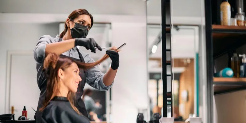 Foto mostra uma mulher sentada em uma cadeira de salão de beleza enquanto outra mulher corta seu cabelo.