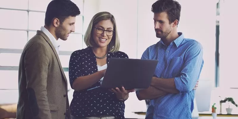 Mulher loira em pé com um computador nas mãos. Ao lado dela estão dois homens. Ela está sorrindo mostrando algo para eles no desktop.