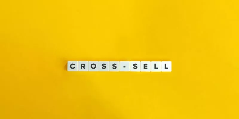 Quadrados brancos e pequenos formando o termo "Cross-Sell" em um fundo amarelo.