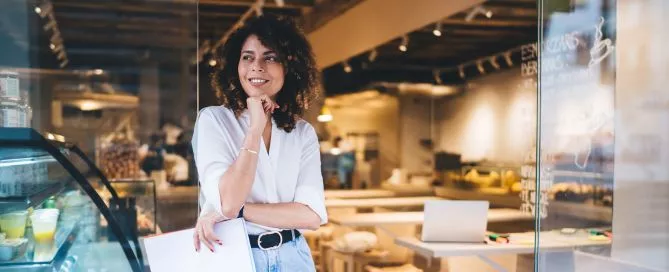 Mulher em frente a um estabelecimento com um braço estendido na cintura e apoiando o maxilar no outro. Ela parece pensativa, mas com um olhar promissor. Representa uma gestora considerando aceitar uma boa proposta de negócio, como uma parceria White Label.