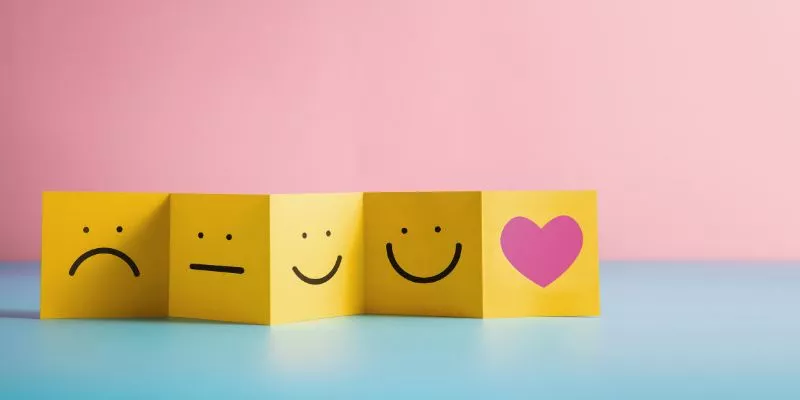 Pequenos quadrados amarelos com rostos desenhados. Da esquerda para a direita, eles vão abrindo mais e mais um sorriso, e o último é um coração rosa.