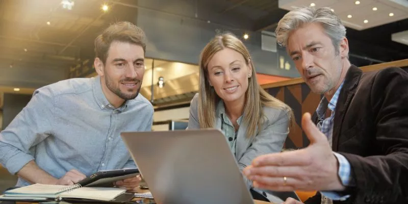 Três pessoas sentadas, enquanto uma delas realiza um pitch de vendas para as outras duas. Elas estão em frente a um computador.