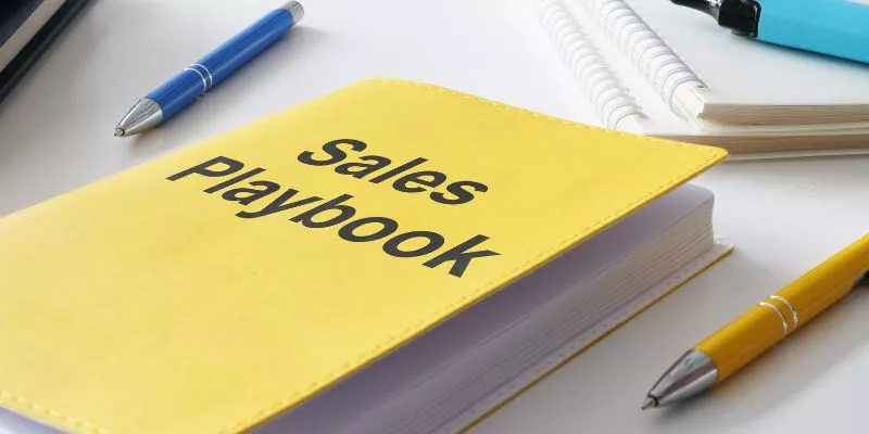 Caderno amarelo em cima de uma mesa. Na capa do caderno está escrito "Sales Playbook", Playbook de Vendas ou Caderno de Vendas em inglês.