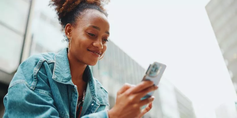 Mulher sorrindo ao ar livre e segurando um smartphone em sua mão direita. Ao que tudo indica, ela está tendo uma experiência Mobile Friendly em seu dispositivo móvel,