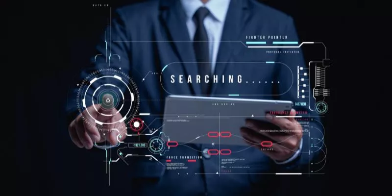 Homem de terno segurando um tablet com um painel digital escrito "searching" acima do dispositivo, simulando uma pesquisa nos mecanismos de busca.