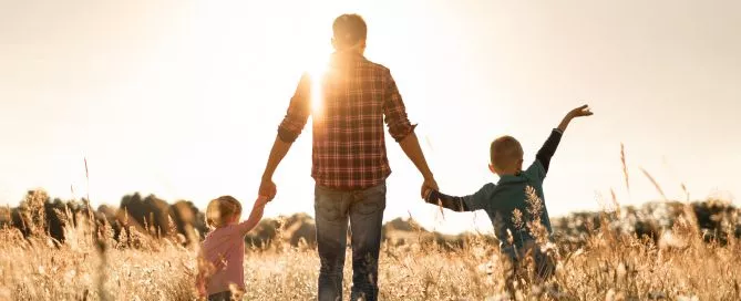 Um pai segurando as mãos de seus dois filhos em um campo de trigo.