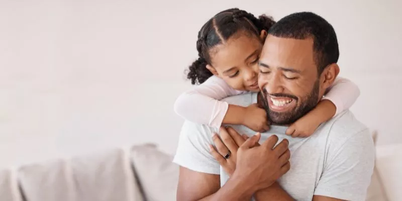 Pai abraçando a filha, que está em suas costas. Ambos estão sorrindo.