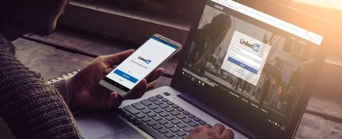 Pessoa navegando pelo app do LinkedIn no celular e de frente para um computador com o site do LinkedIn aberto.
