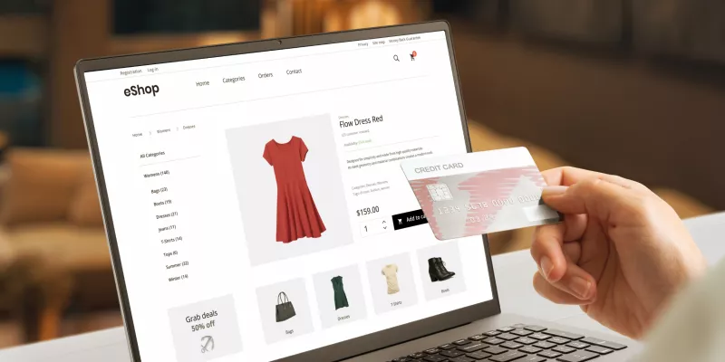A foto mostra a tela de um notebook em foco onde aparece a tela de uma loja online. Em primeiro plano tem a mão de uma pessoa segurando um cartão de crédito.