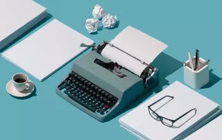 A foto possui um fundo azul que simula uma mesa onde, no meio, está uma maquina de escrever também azul e ao lado direito e esquerdo estão pilhas de papéis. E uma das pilhas tem um óculos.