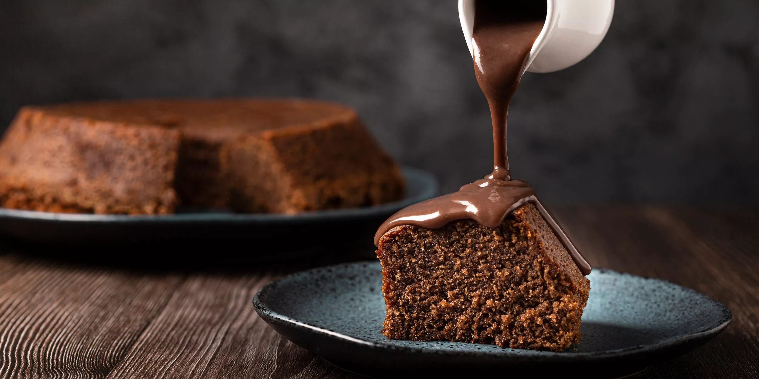 A imagem mostra um pedaço de bolo de chocolate em primeiro plano, logo atrás está o bolo completo. Acima do pedaço de bolo tem um recipiente derramado calda de chocolate.