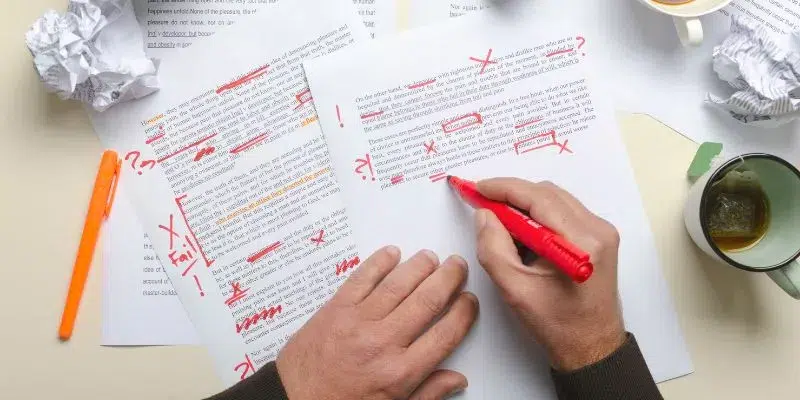 A imagem mostra a mão de um homem segurando uma caneta vermelha, em uma mesa estão algumas folhas onde esse homem anotou algumas informações.