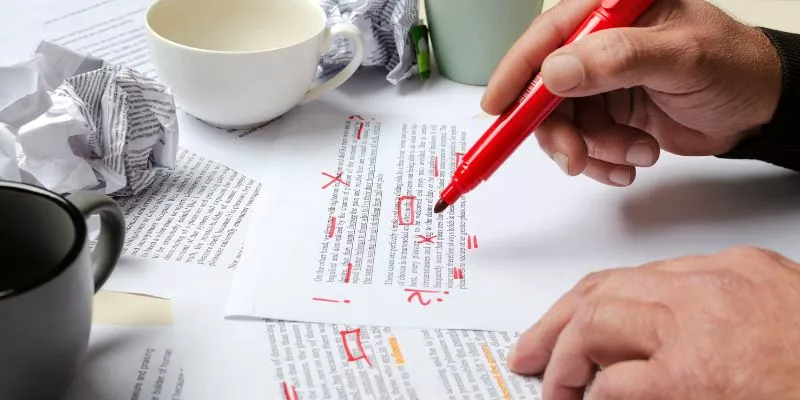 A imagem mostra a mão de um homem segurando uma caneta vermelha, em uma mesa estão algumas folhas onde esse homem anotou algumas informações.